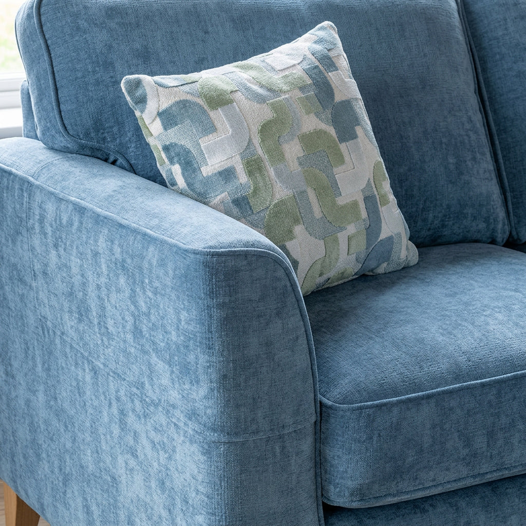 A close-up of a blue upholstered sofa with an arm cap featuring a white, sage green, and blue geometric pattern cushion.