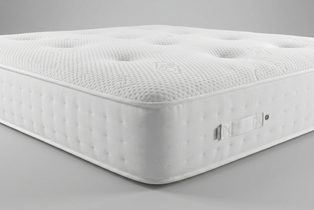 Close-up of a mattress corner showing the white textured top surface with tufted dots and the gray side panel, with part of a blue patterned blanket and pillows visible in the background.