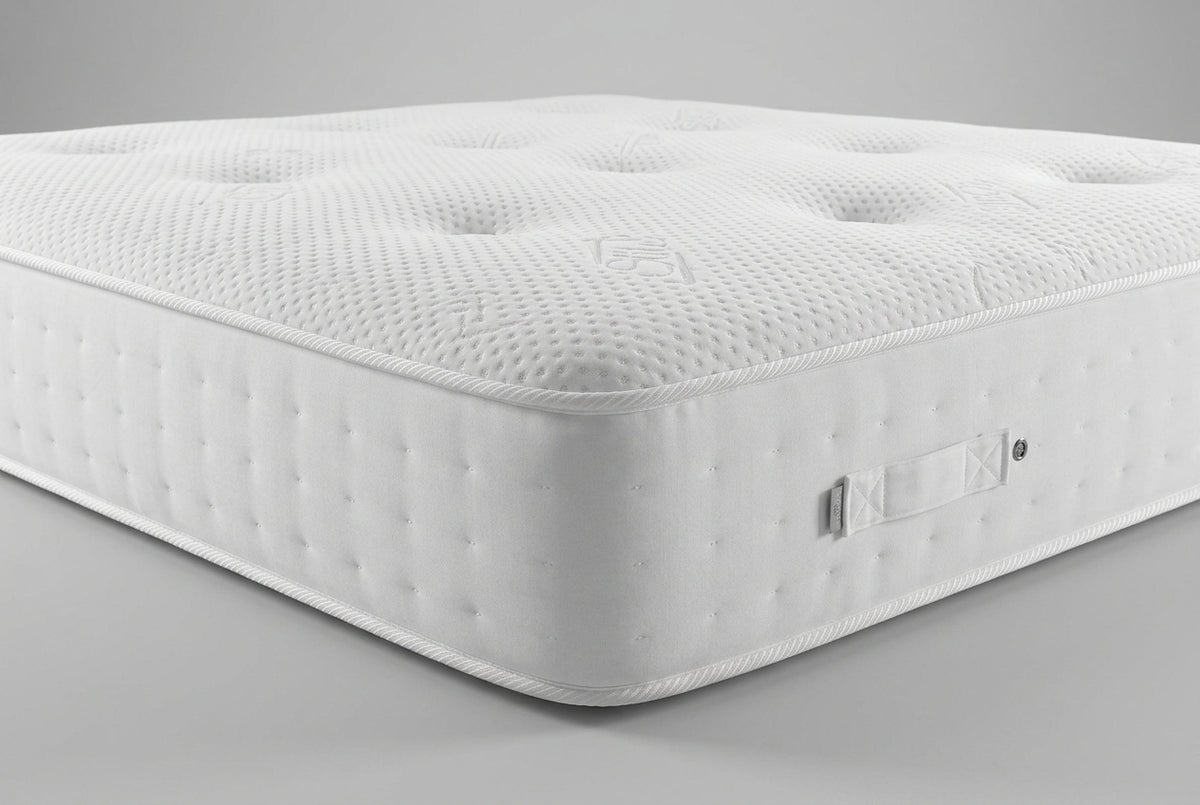 Close-up of a mattress corner showing the white textured top surface with tufted dots and the gray side panel, with part of a blue patterned blanket and pillows visible in the background.