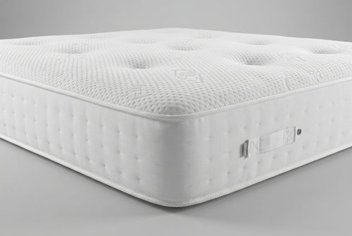 Close-up of a mattress corner showing the white textured top surface with tufted dots and the gray side panel, with part of a blue patterned blanket and pillows visible in the background.