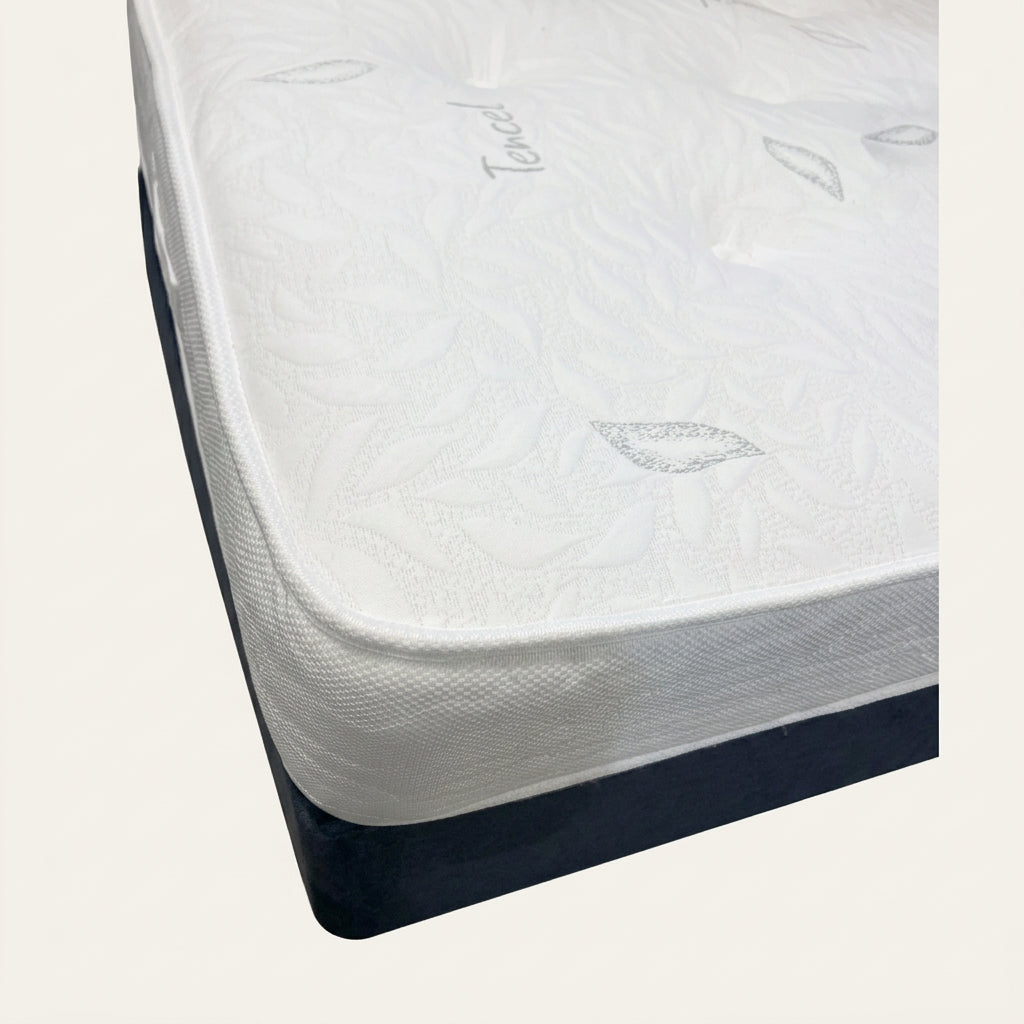 A close-up of a white Tencel mattress showing its textured, tufted top surface with a leaf pattern and the brand name 'Tencel' embroidered on it, resting on a dark blue bed frame.